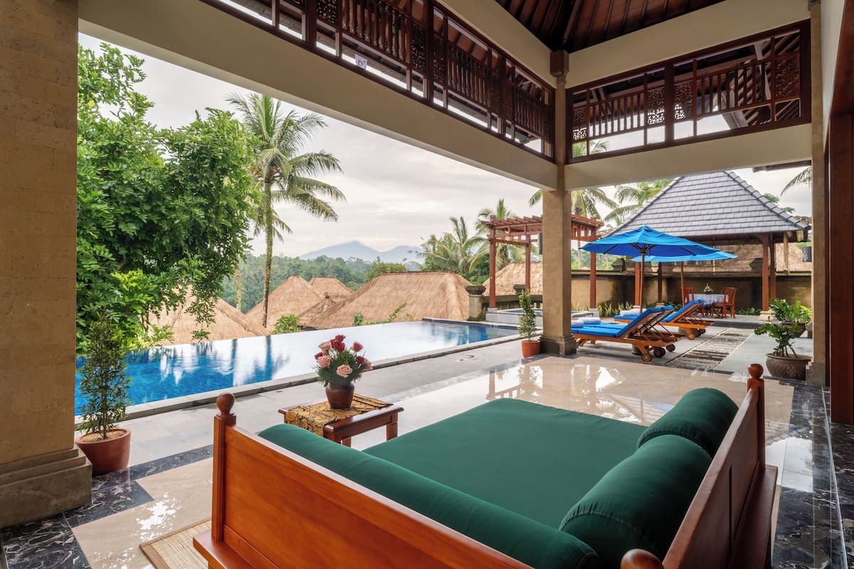 A room with a bed, a view of a swimming pool, and a lounge area with umbrellas.