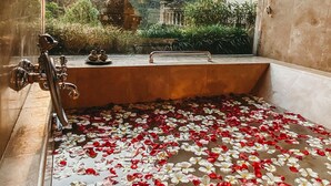 A bathtub filled with water and rose petals, with a view of a garden and a stone structure outside. - Puri Wulandari a Boutique Resort and Spa (Ubud)