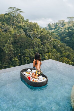 Une personne assise sur une plateforme flottante dans une piscine, avec un panier rempli de nourriture et de boissons, entourée d’une végétation luxuriante.