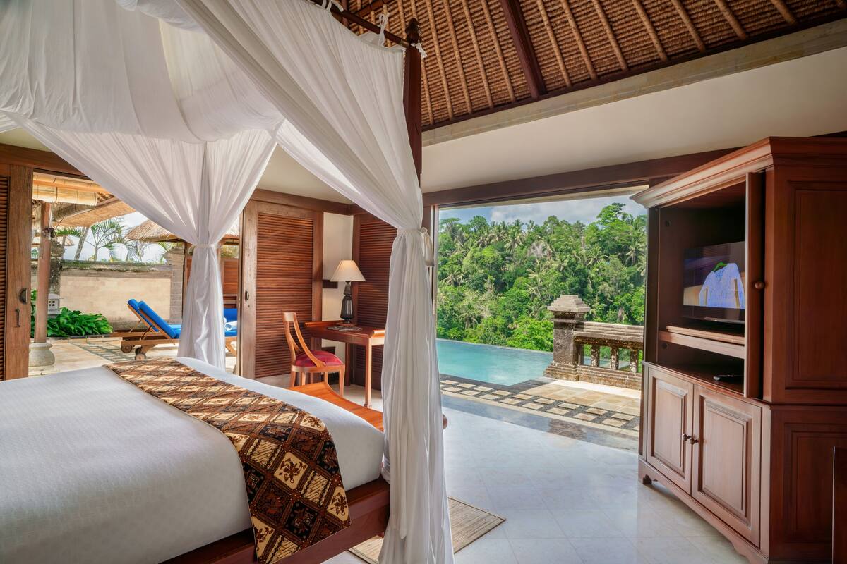 A bedroom with a bed, a TV, and a view of a swimming pool and lush greenery.
