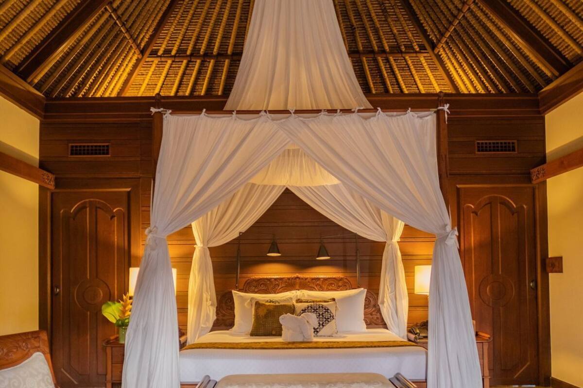 A bedroom with a four-poster bed, a wooden bench, and a table with fruit and a statue.