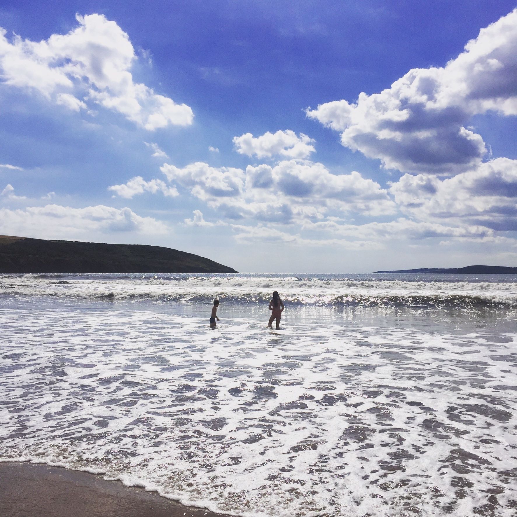 Uma praia com ondas, céu claro e colinas ao longe.