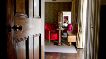 Una puerta de madera con manijas de latón, una butaca tapizada de rojo, un sillón color beige, una mesa redonda con un florero y un espejo en la pared.