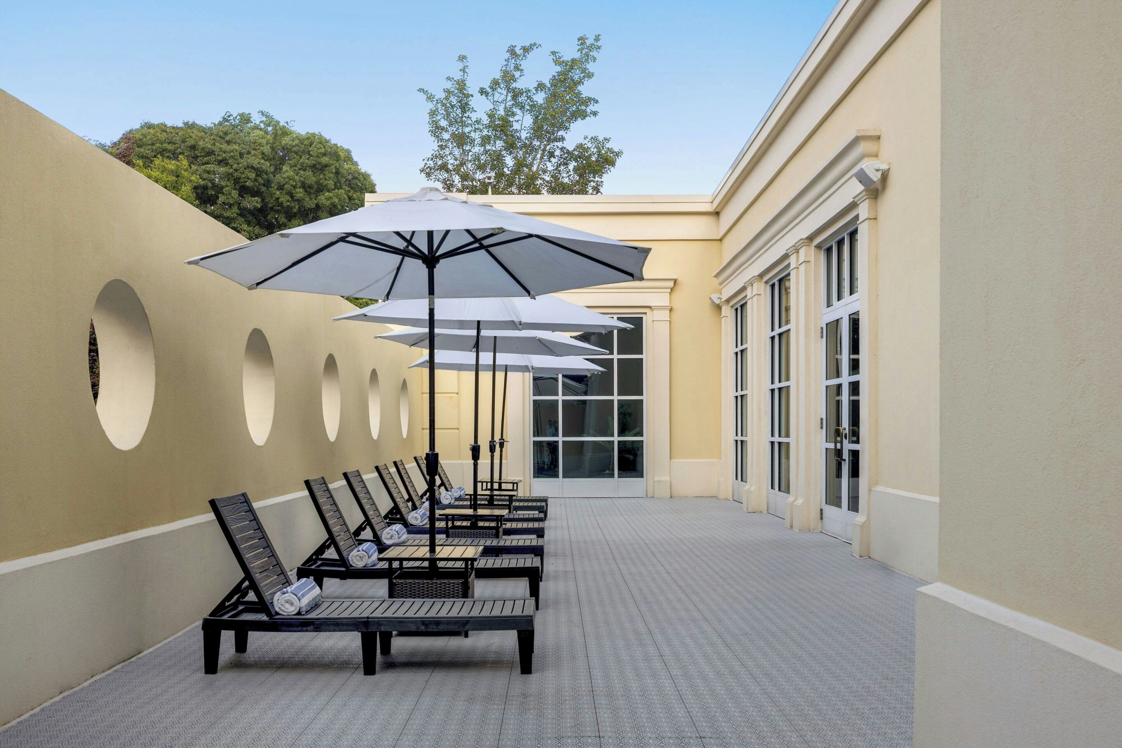 A covered outdoor seating area with lounge chairs and umbrellas, a tiled floor, and a building with large windows and a beige wall.