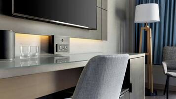A modern hotel room with a flat-screen TV, a glass desk, a lamp, and two chairs.