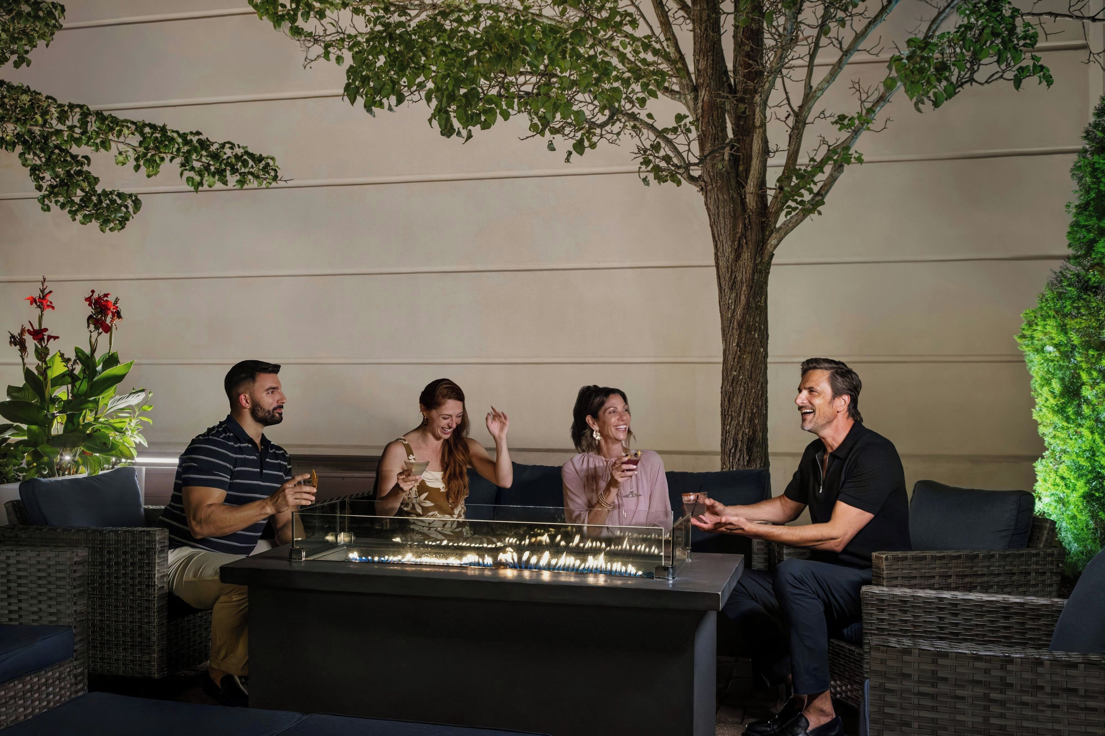 A group of people seated around a bar area with a fire feature, wicker furniture, and indoor plants.