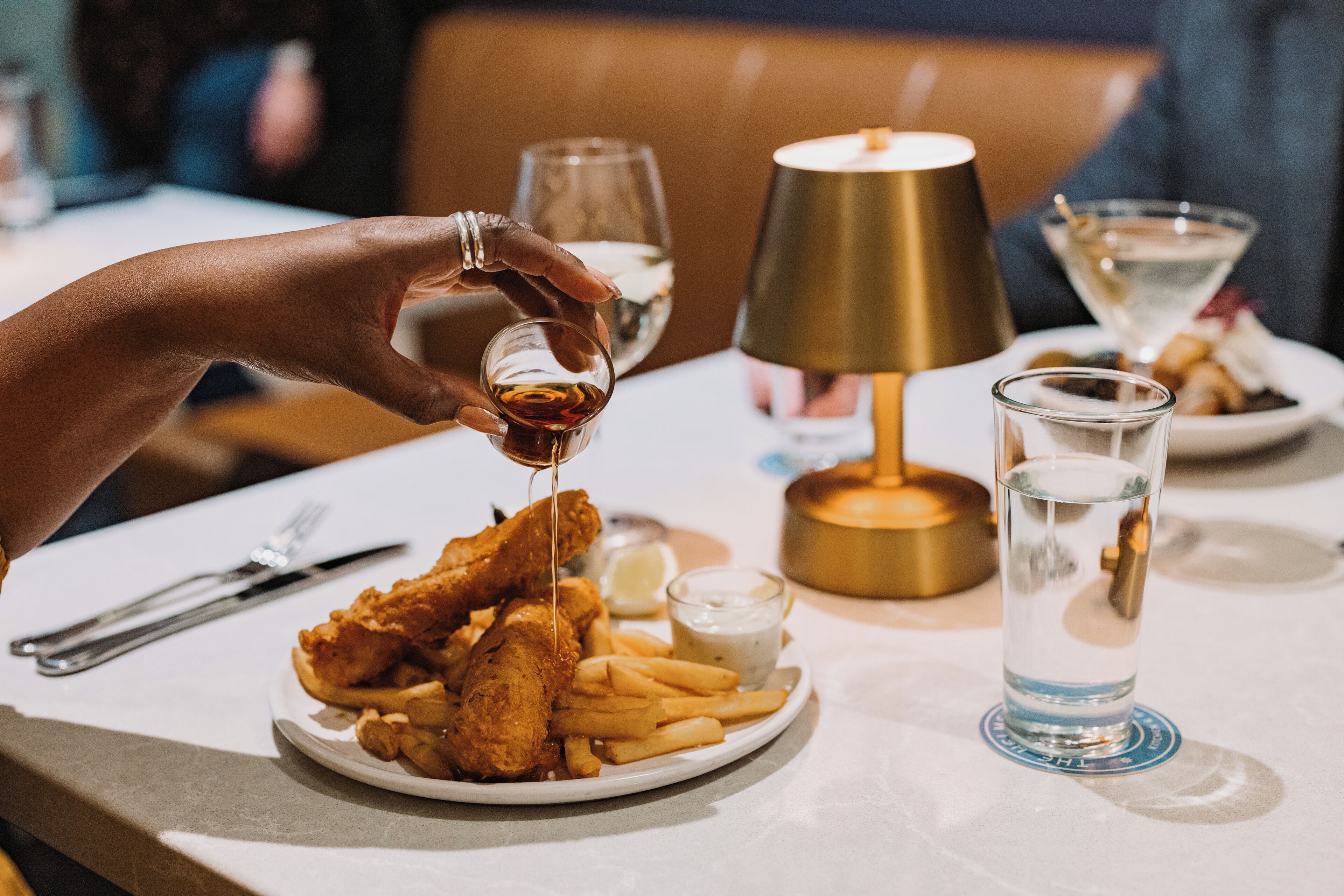 Un plato de pescado con papas fritas, un vaso de agua y una lámpara sobre una mesa.