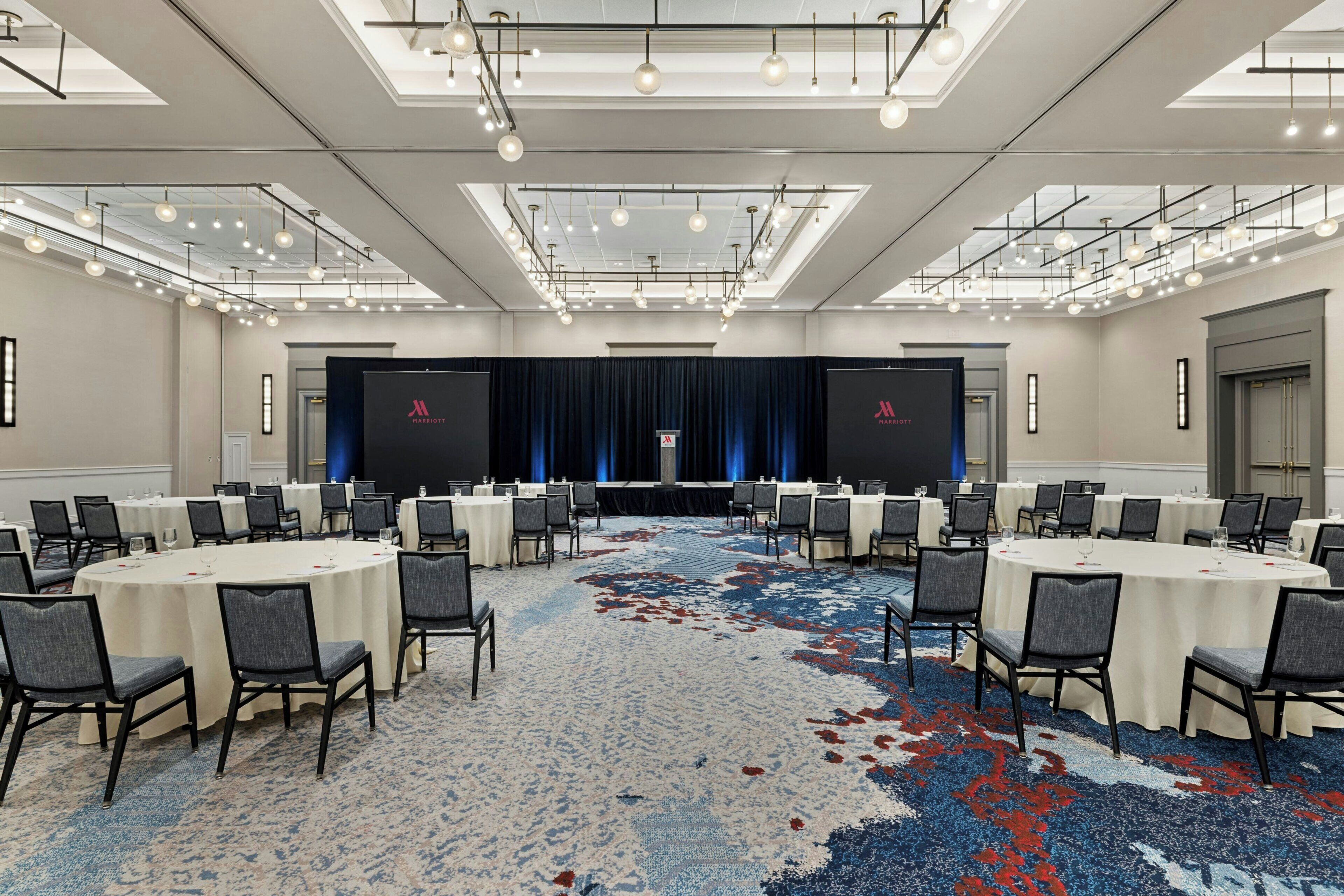 A conference room with round tables, chairs, a stage with curtains, and a patterned carpet.