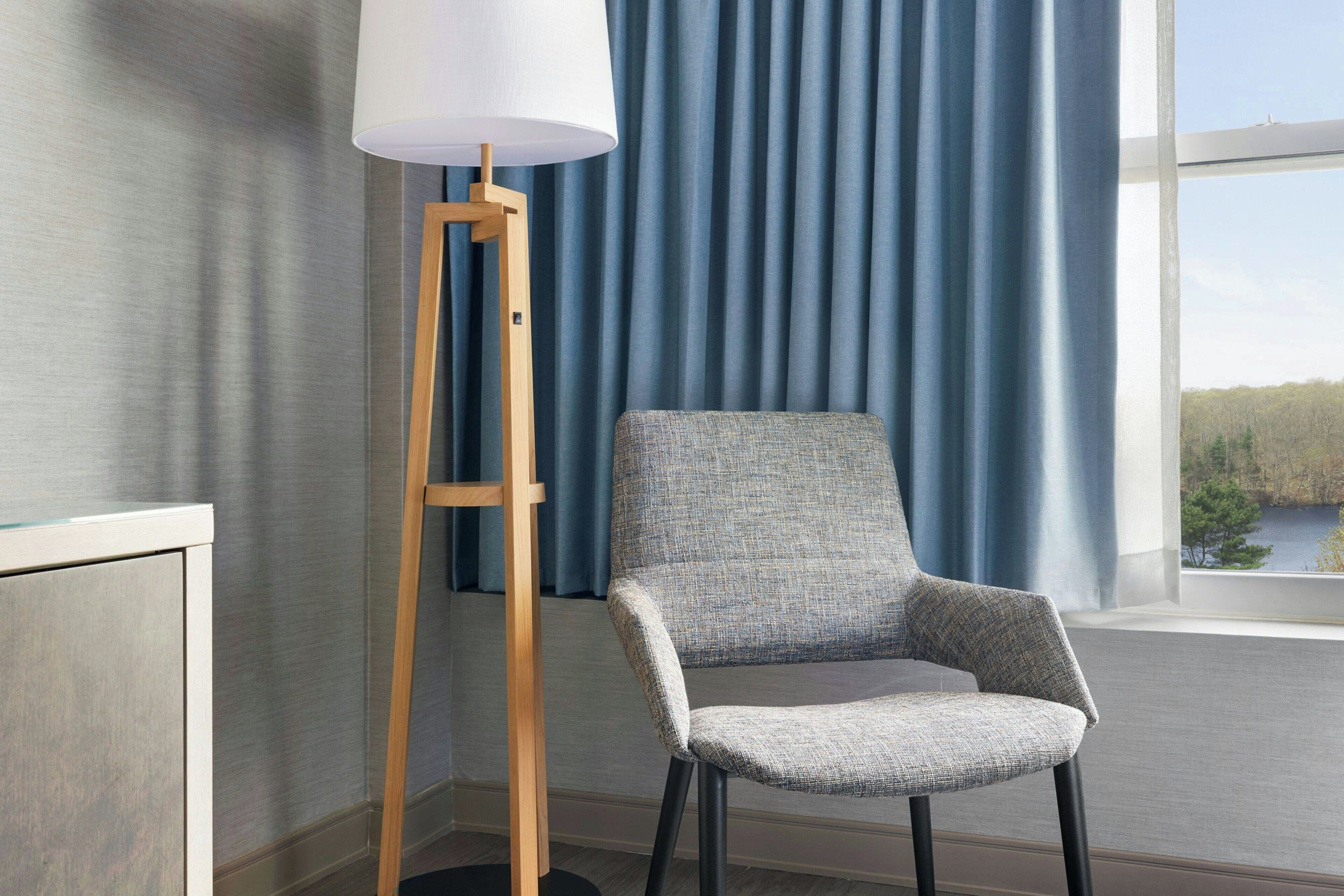 Een kamer met een hoge vloerlamp, een grijs gestoffeerde fauteuil en uitzicht op een wateroppervlak door een raam met blauwe gordijnen.