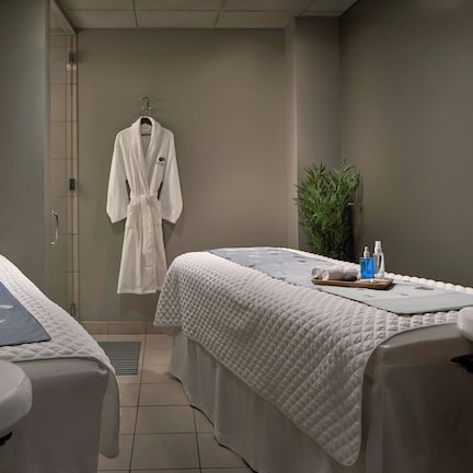 A spa room with two massage tables, a robe hanging on the wall, and a plant.