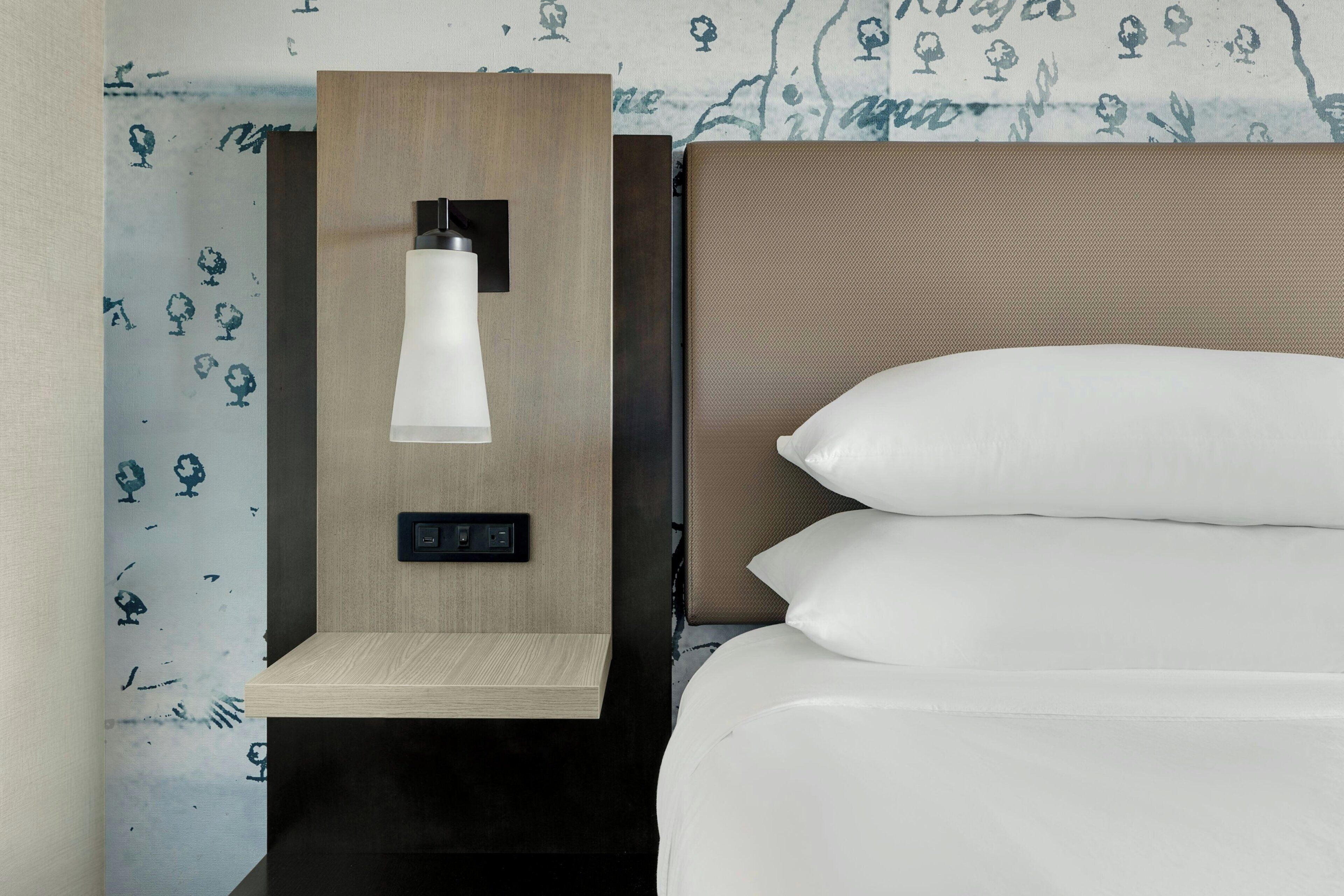 Una habitación de hotel con una cama, una mesita de noche con una lámpara y un papel tapiz con motivos florales.