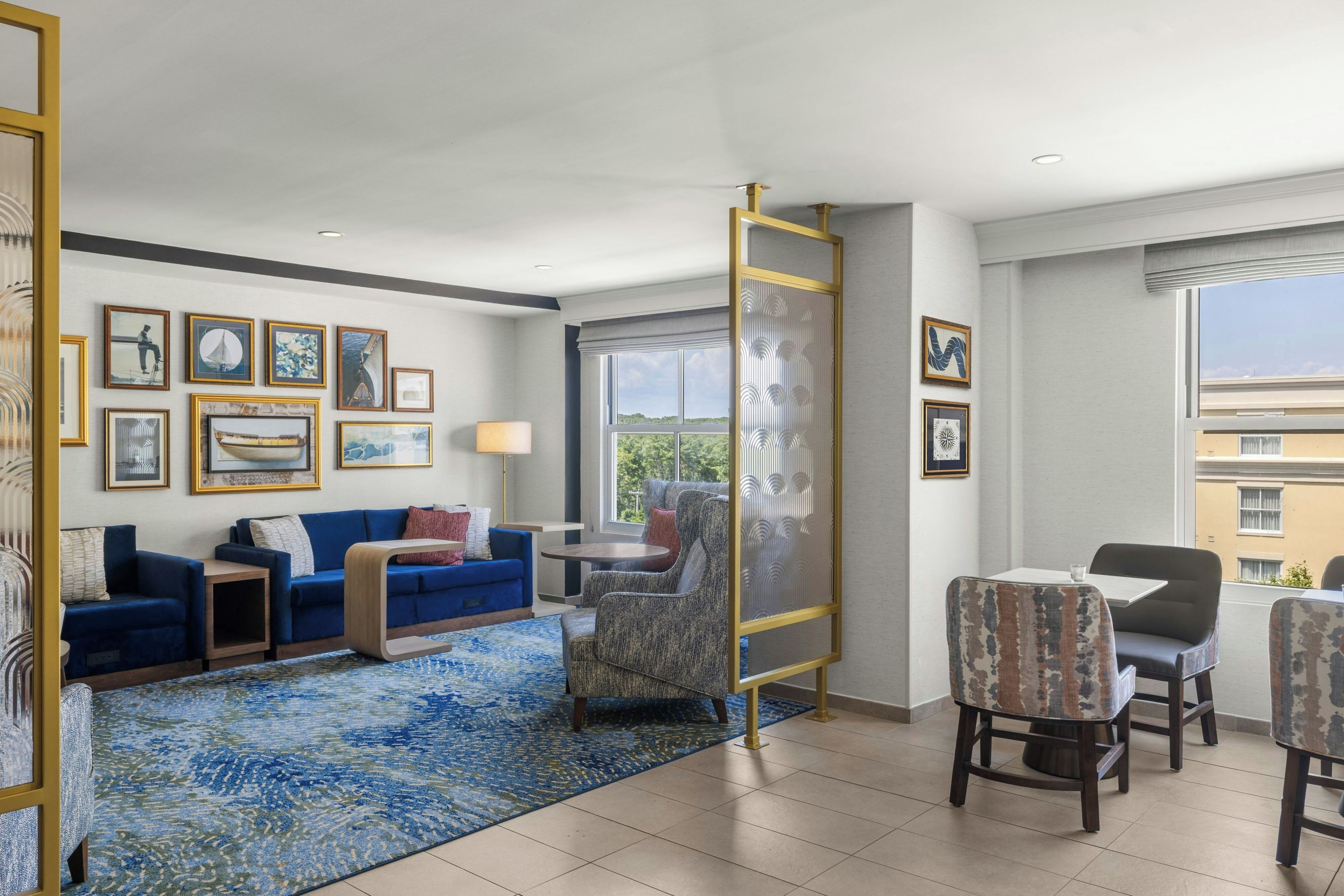 A hotel room with a blue sofa, a dining table with chairs, a large window with a view, and various framed artworks on the walls.