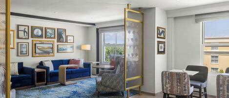 A hotel room with a blue sofa, a dining table with chairs, a large window with a view, and various framed artworks on the walls.