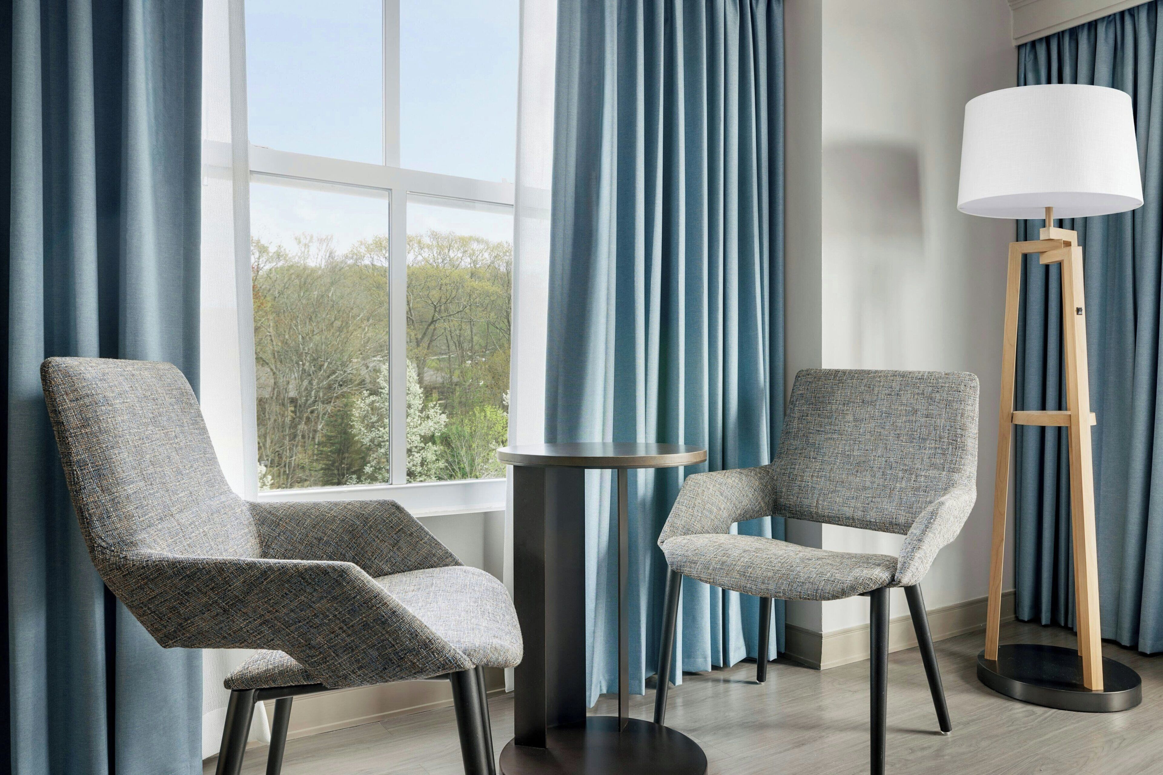 A room with two grey upholstered chairs, a round table, a floor lamp, and a view through a window with blue curtains.