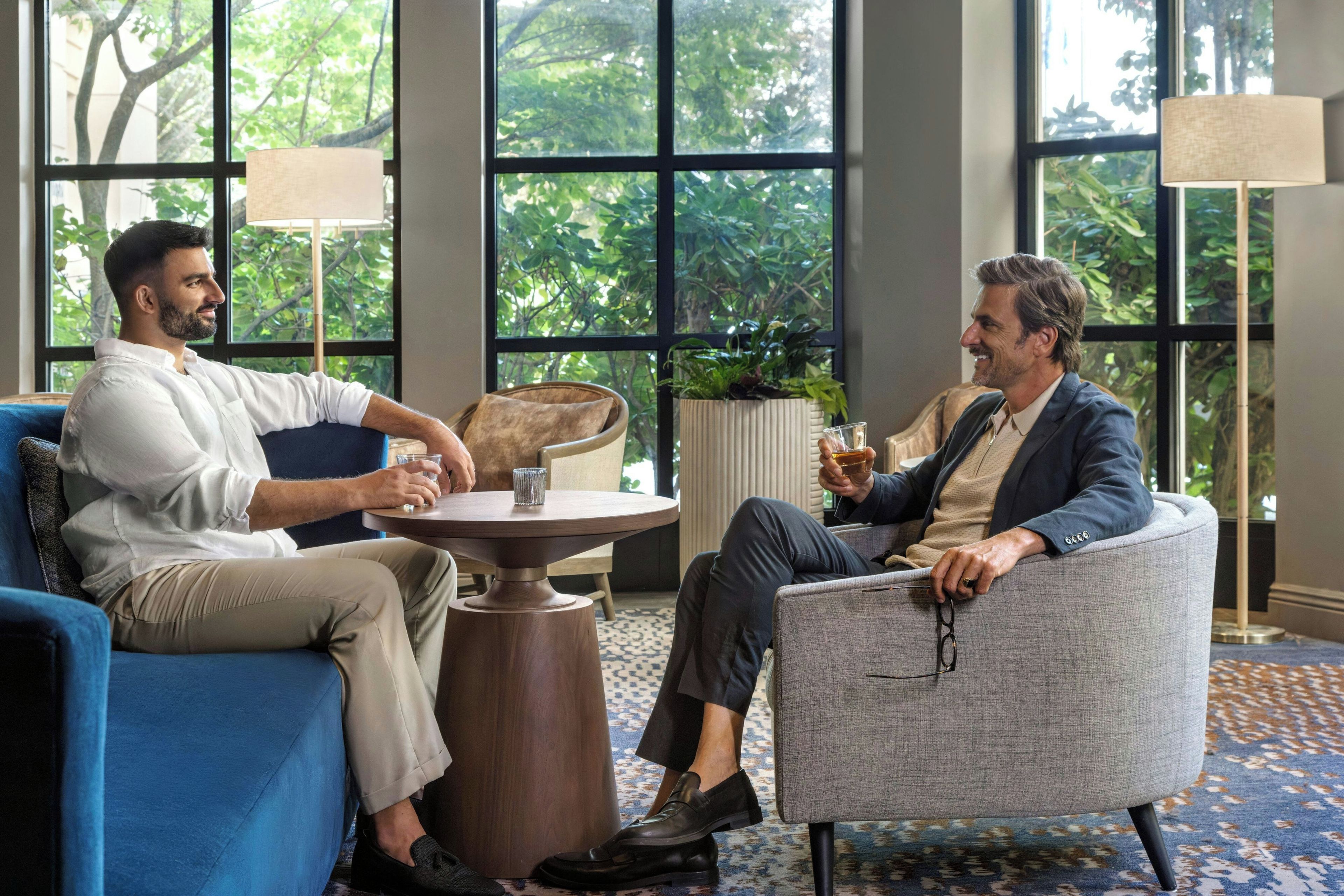 Two individuals seated in a modern lounge area with a round table, large windows, and a view of greenery outside.