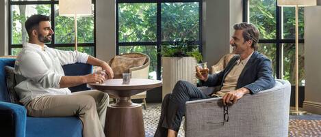 Two individuals seated in a modern lounge area with a round table, large windows, and a view of greenery outside.
