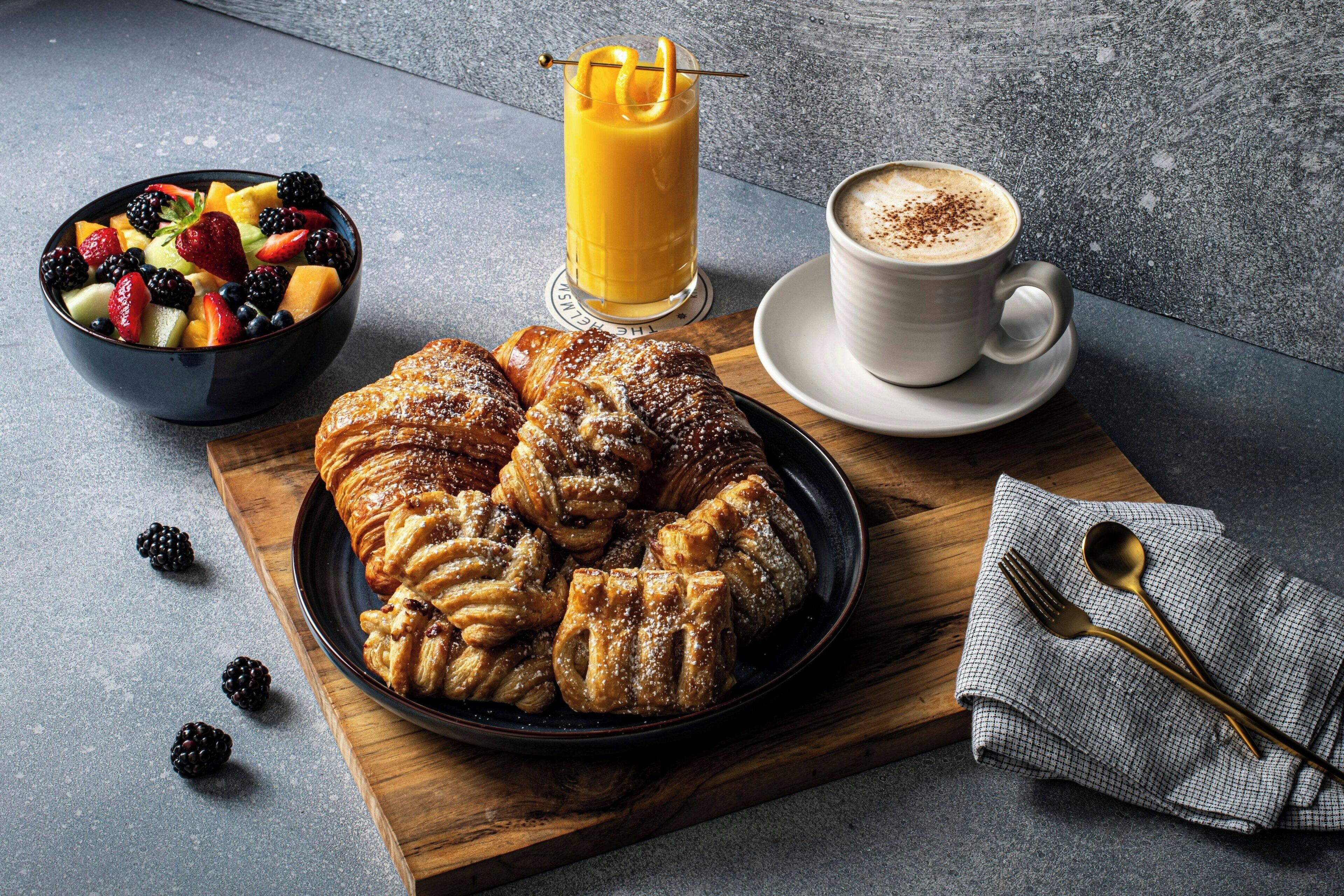 A breakfast setting with pastries, a fruit bowl, a glass of orange juice, and a cup of coffee on a wooden board.