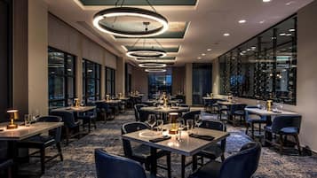 A restaurant with multiple tables and chairs, a ceiling with circular light fixtures, and large windows on one side.