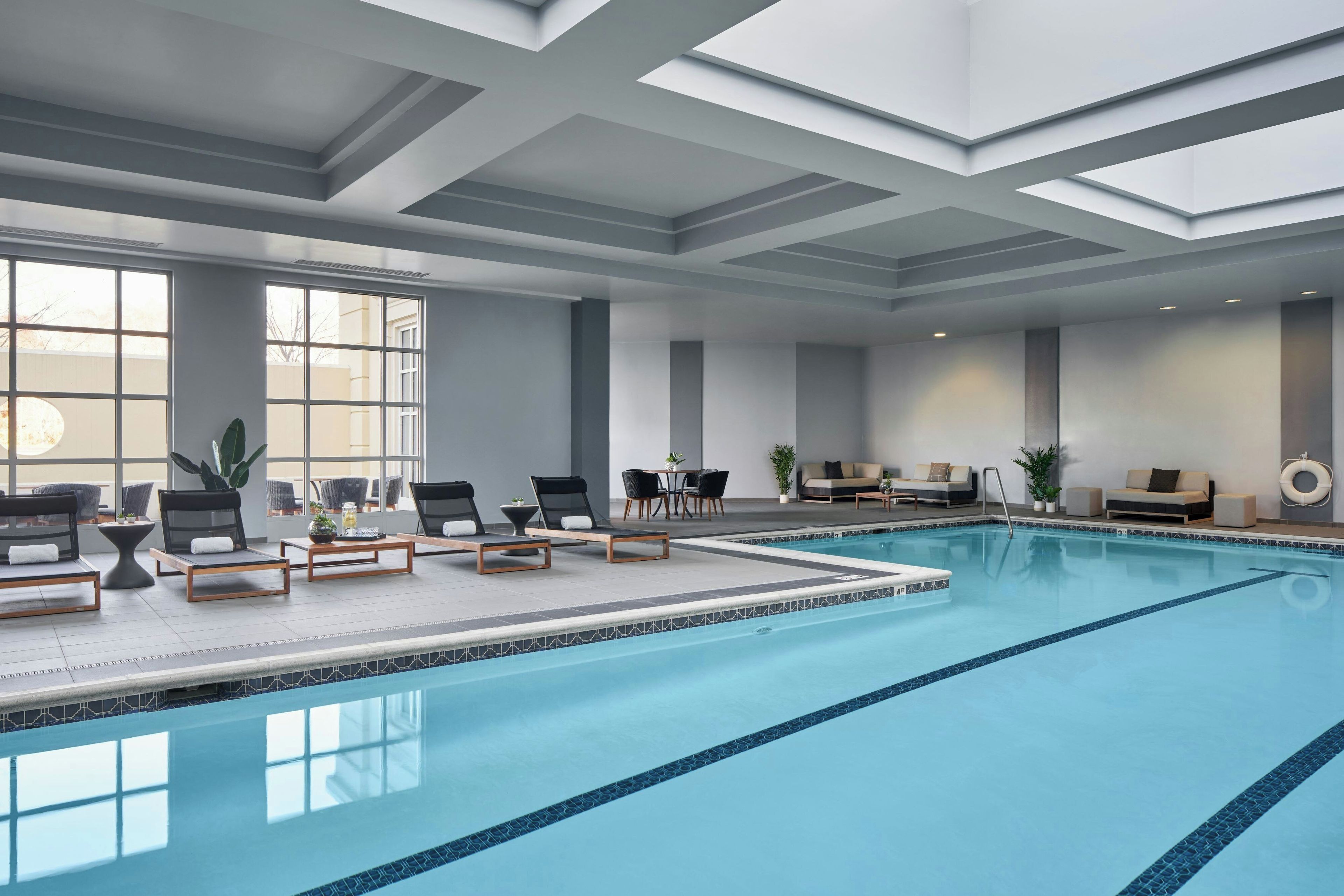 Una piscina interior con sillas de descanso y mesas, ventanas amplias y un diseño moderno en el techo.