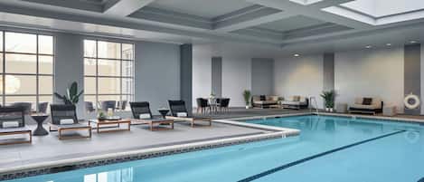 Una piscina interior con sillas de descanso y mesas, ventanas amplias y un diseño moderno en el techo.