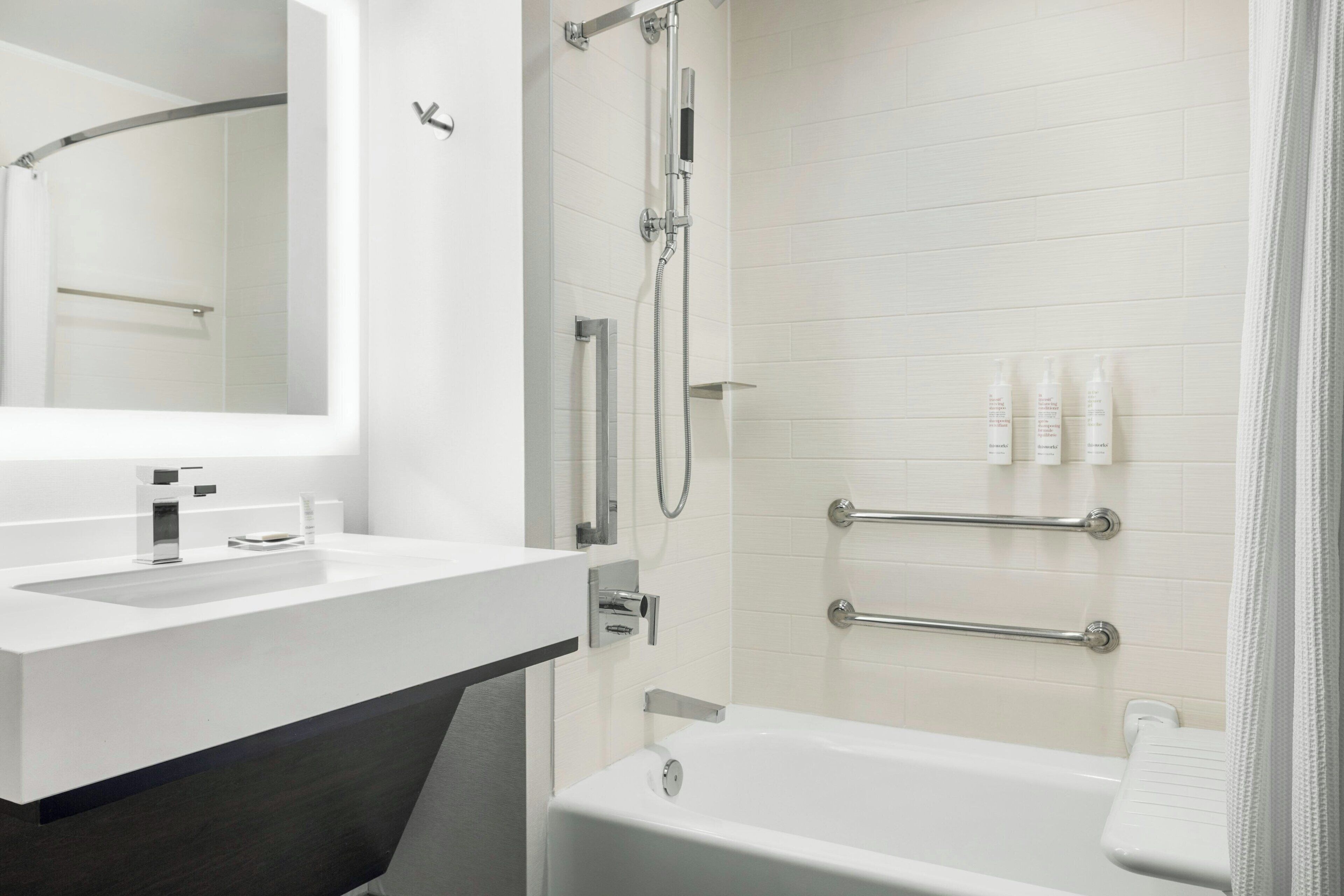 Een moderne badkamer met een witte wastafel, spiegel en bad. Er zijn handgrepen aan de wanden en een douchekop.