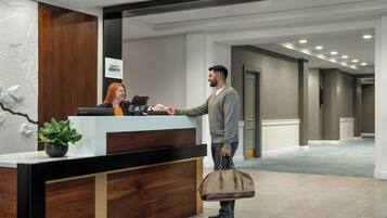 Mostrador de recepción de hotel con una recepcionista, un huésped de pie y una planta en la barra.