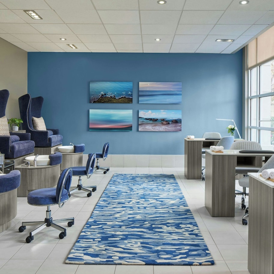 A modern spa room with a blue wall, four framed artworks, a blue patterned rug, and a reception desk with a computer and a plant.