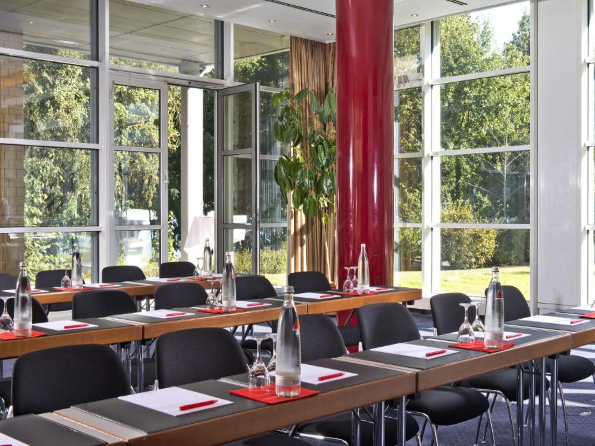 Ein Konferenzraum mit mehreren Tischen, Stühlen und einem großen Fenster mit Blick auf Bäume.