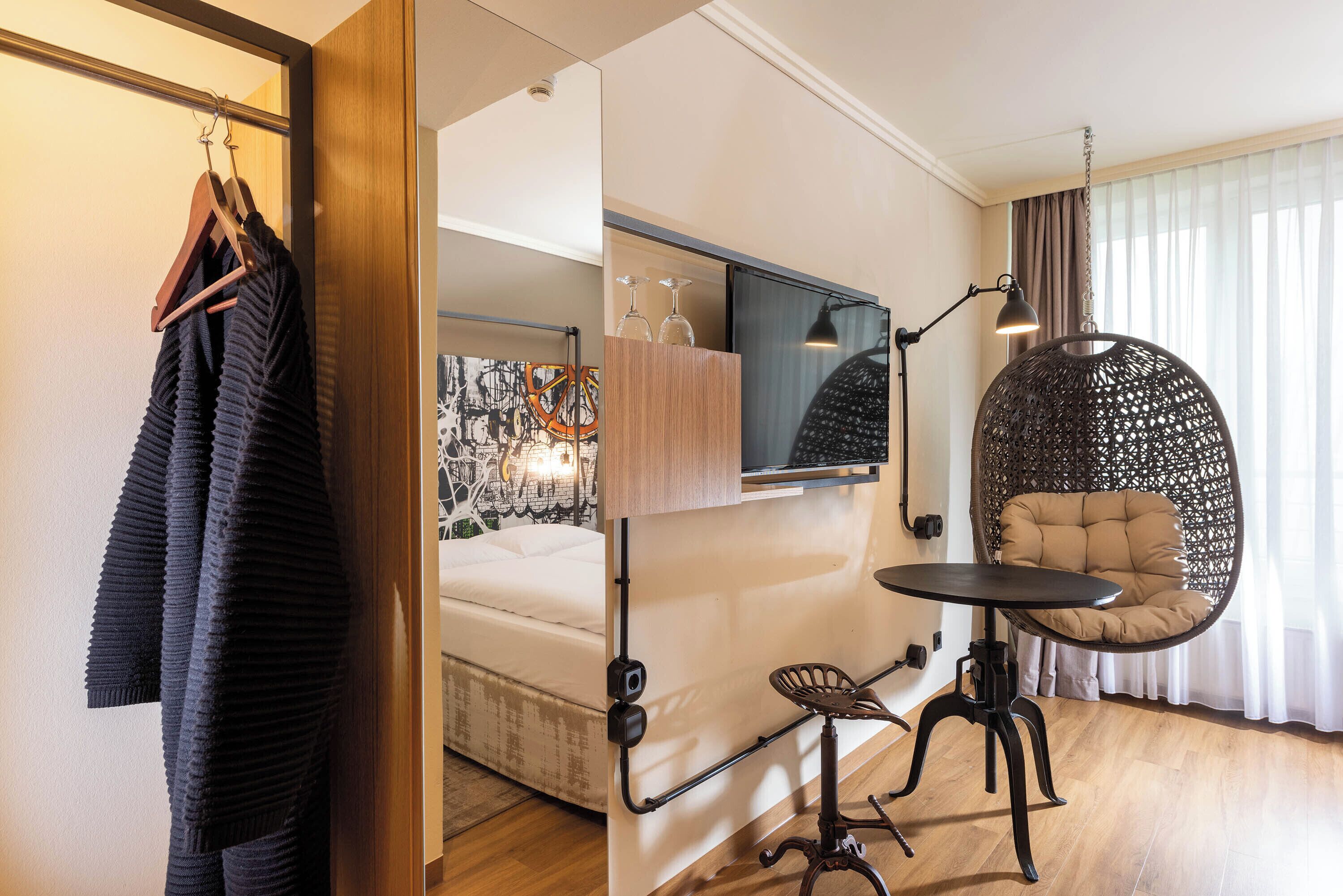 A compact hotel room with a bed, a small table, a hanging chair, and a TV mounted on the wall.