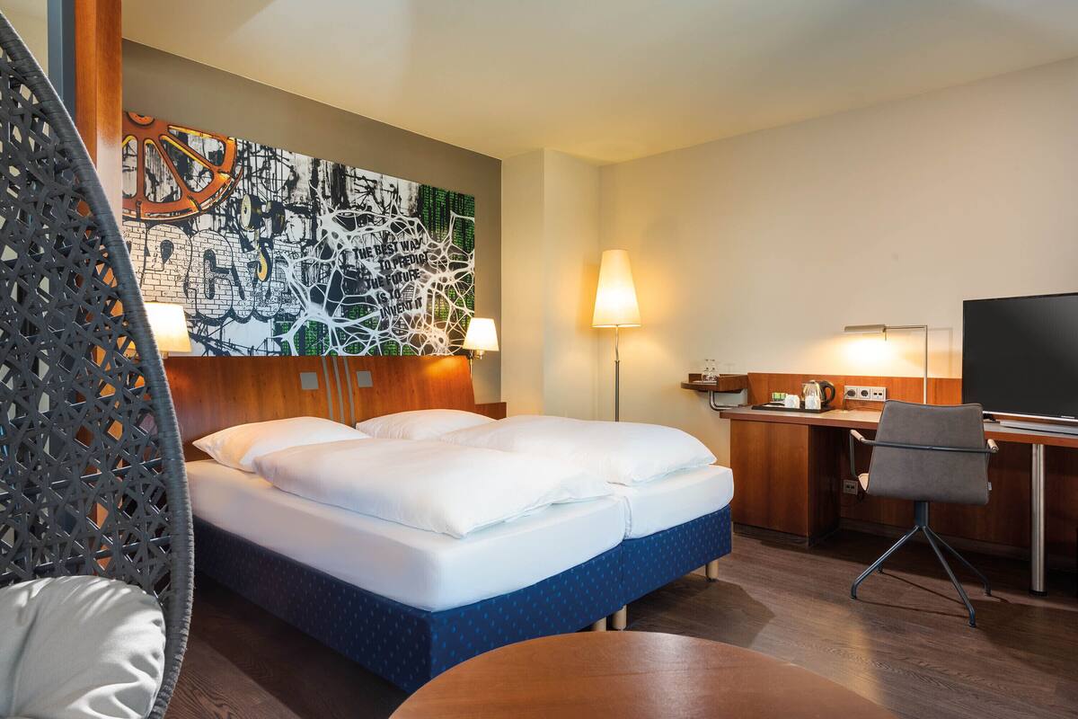 A hotel room with a bed, desk, chair, and a wall with a large abstract painting.