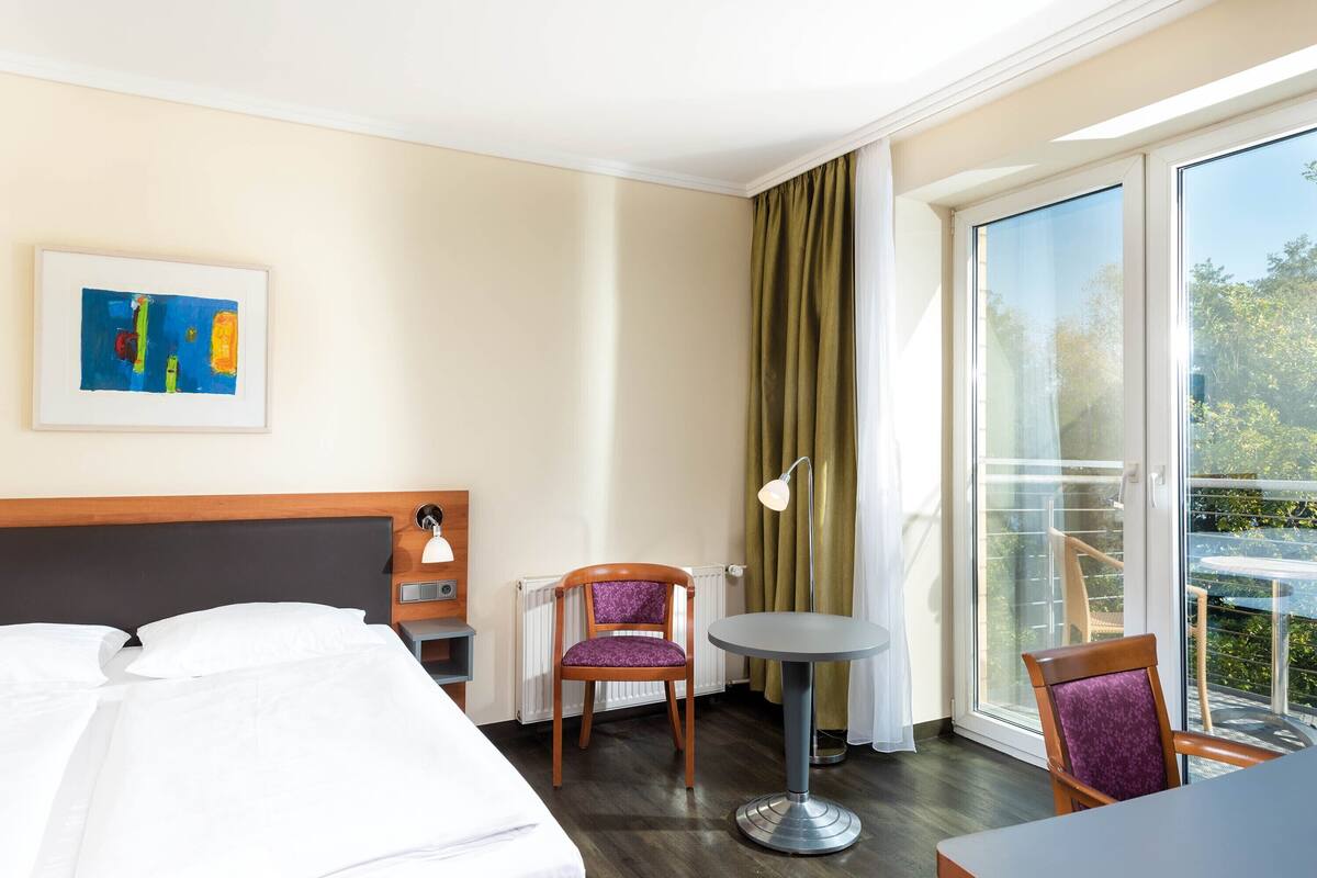 A hotel room with a bed, a chair, a small table, a painting, a lamp, and a view of the outdoors.
