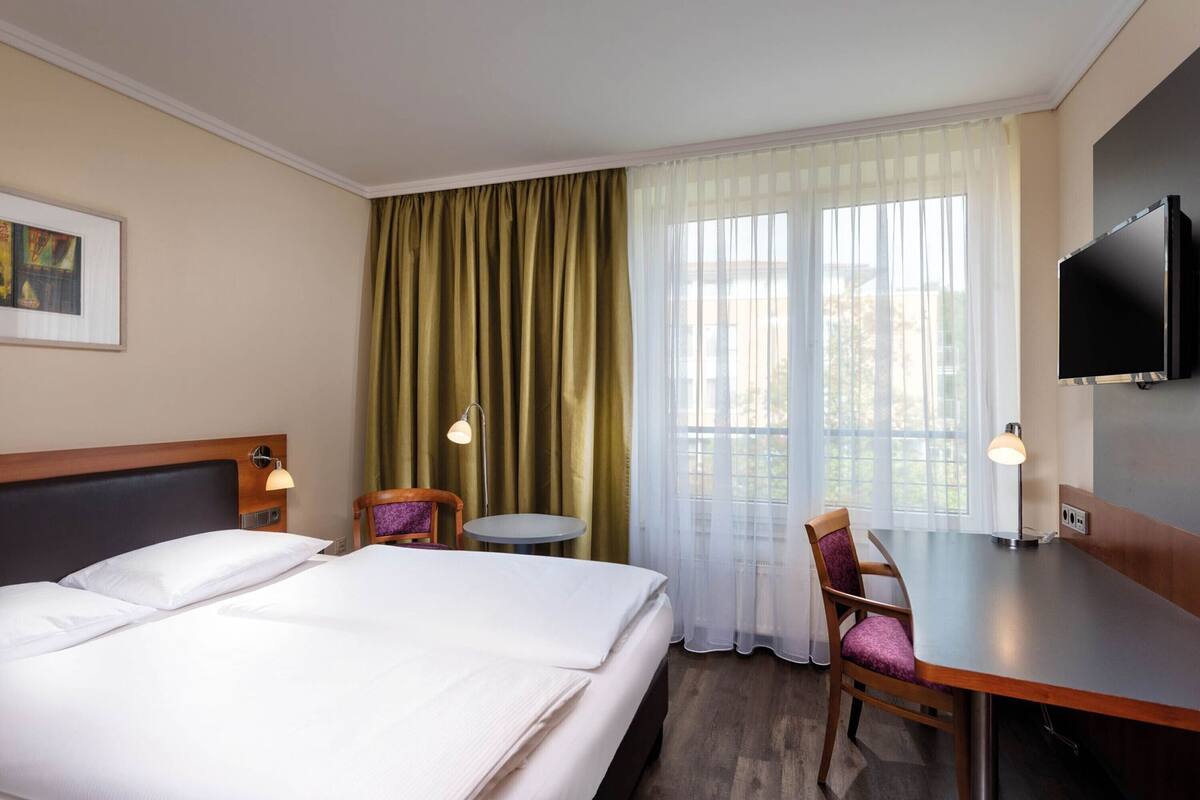 A hotel room with a bed, two chairs, a desk, a television, and a window with curtains.