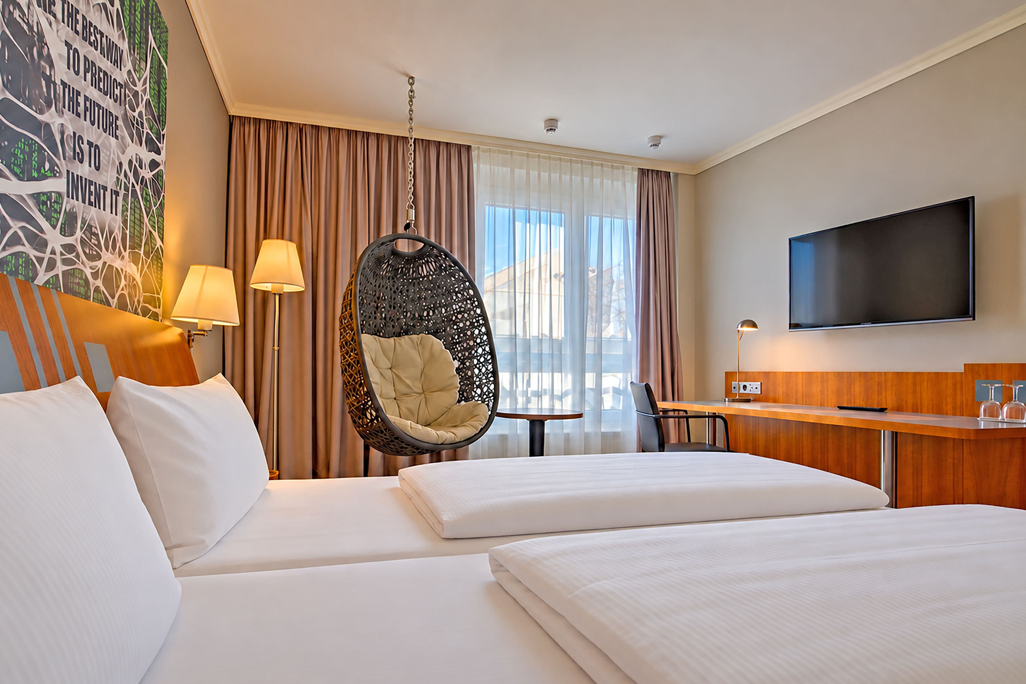 A hotel room with a bed, a hanging chair, a desk, a television, and a window with curtains.