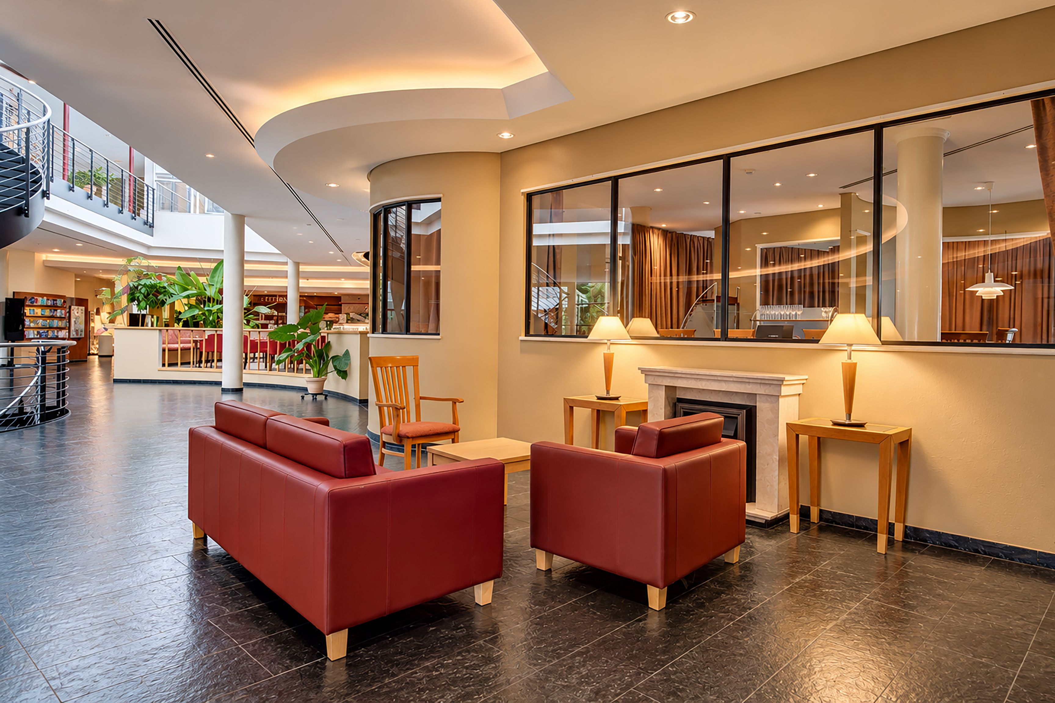 Eine moderne Lobby mit roten Ledersofas, einem Kamin und einem großen Fenster, das den Innenraum spiegelt.