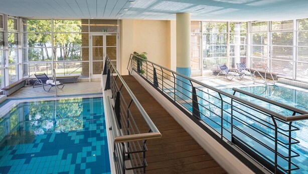 An indoor swimming pool with a wooden deck, glass railings, and lounge chairs. - Seminaris Seehotel Potsdam (Potsdam)