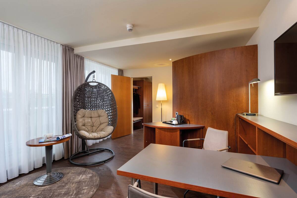 A modern hotel room with a wooden desk, a laptop, a hanging chair, a small table with a magazine, and a lamp.