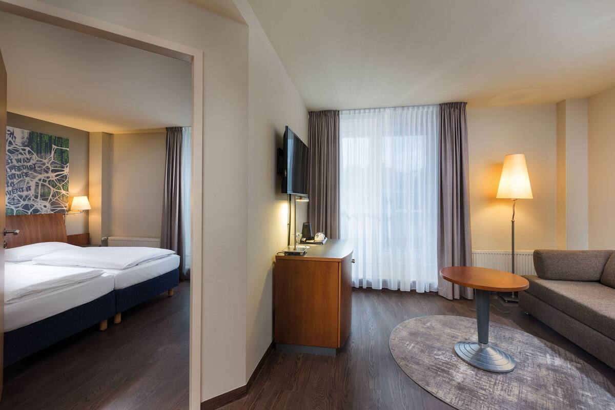 A hotel room with a bed, a desk with a lamp, a TV, a sofa, and a window with curtains.