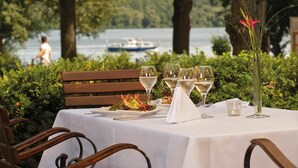 An outdoor dining table with a white tablecloth, surrounded by greenery, with a lake and a boat in the background. - Seminaris Seehotel Potsdam (Potsdam)