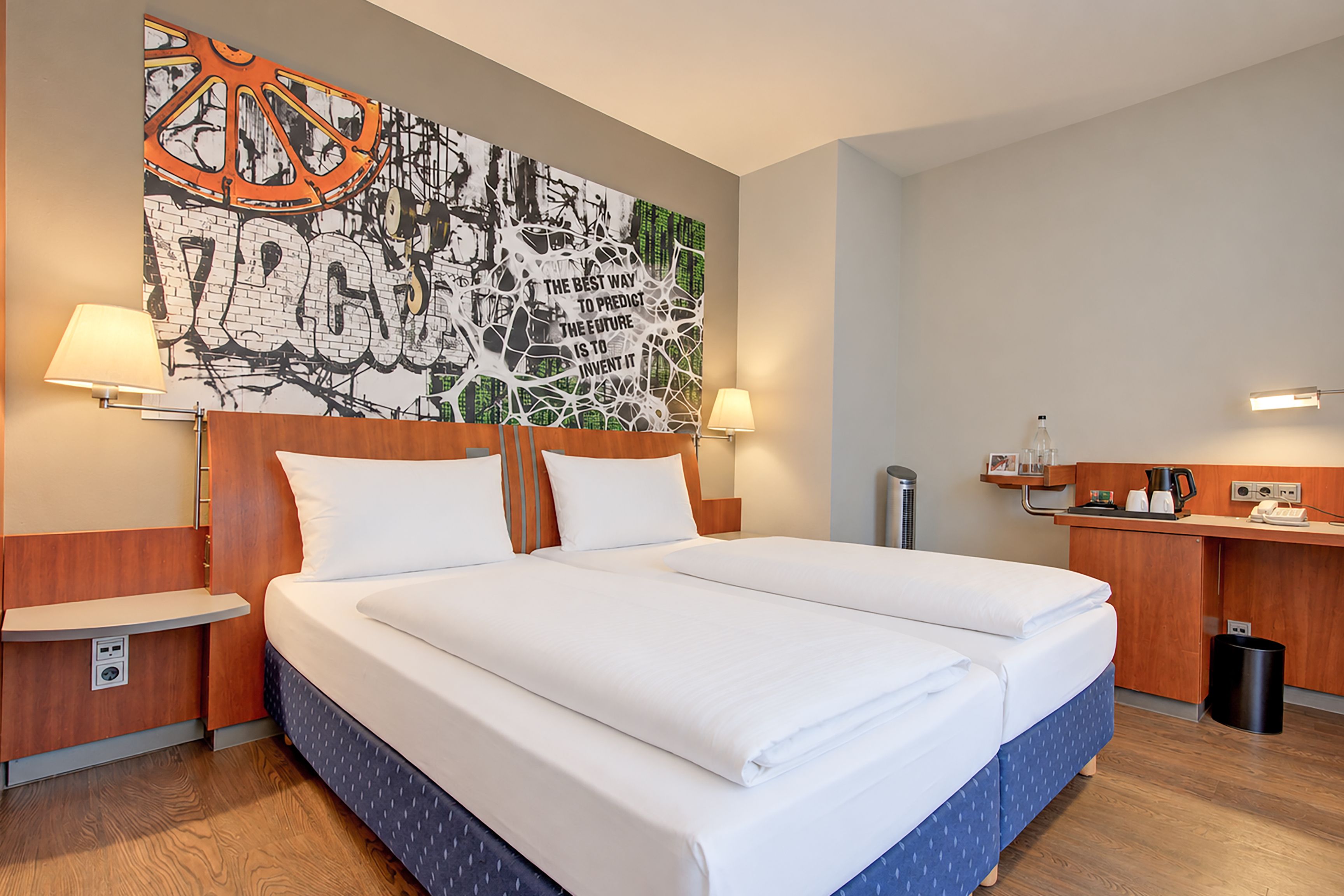 Una habitación de hotel con una cama grande, dos mesitas de noche, una lámpara de pared, un escritorio pequeño con una silla y un cesto de basura.