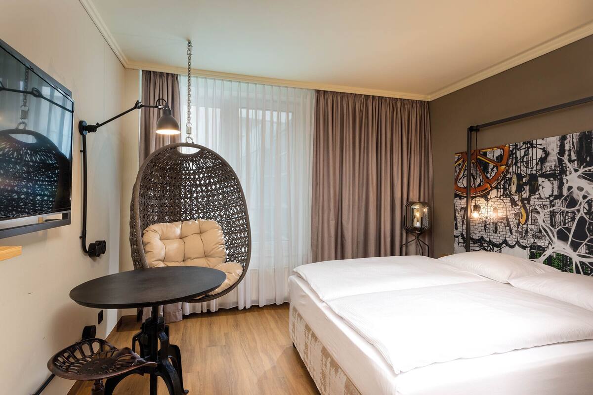A hotel room with a bed, a round chair, a small table, a wall-mounted lamp, and a large abstract painting.