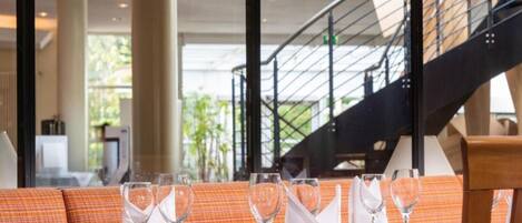 Un ambiente de restaurante con mesas dispuestas para comer, una escalera al fondo y grandes ventanales que dejan entrar luz natural.