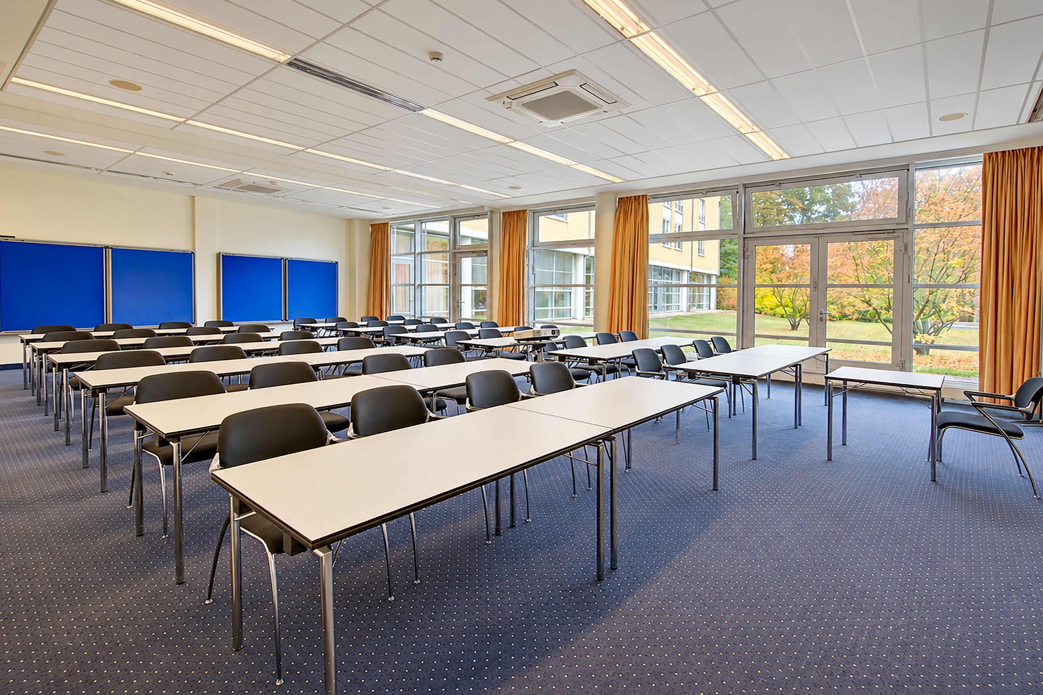 A conference room with multiple tables and chairs, large windows with curtains, and a blue carpet.