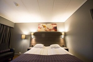 A hotel room with a large bed, two bedside tables, a headboard, a wall-mounted artwork, and a floor lamp.