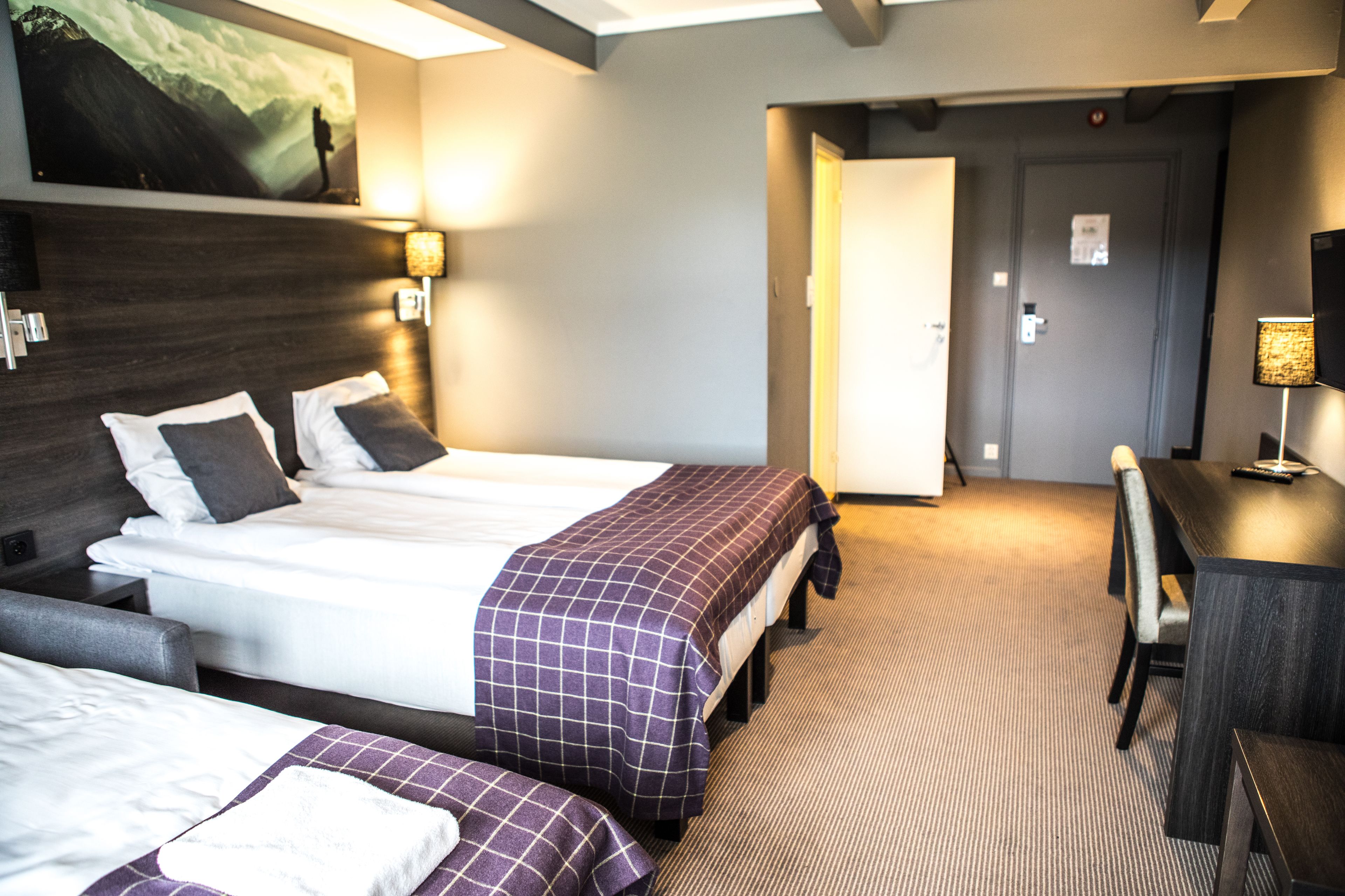 A hotel room with two beds, a desk, and a chair. There is a painting on the wall and a door leading to another room.