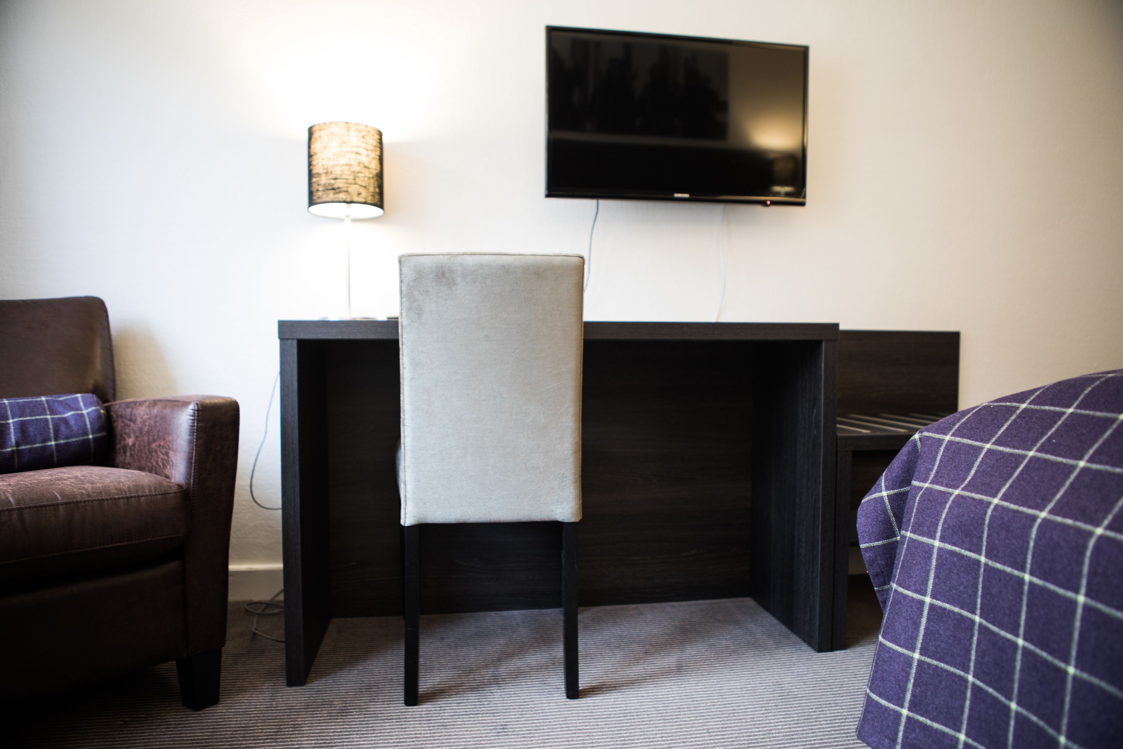 Habitación de hotel con escritorio, silla, televisor montado en la pared y cama con colcha a cuadros.
