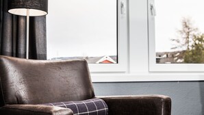 A brown leather armchair with a plaid pillow, positioned near a window with a view of buildings and trees.