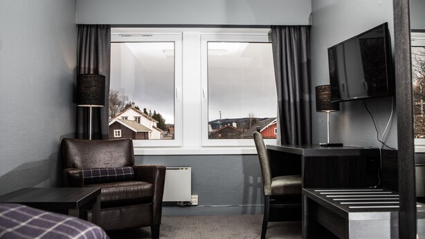 Habitación de hotel con cama, escritorio, dos sillas, televisor y ventana con vista a casas.