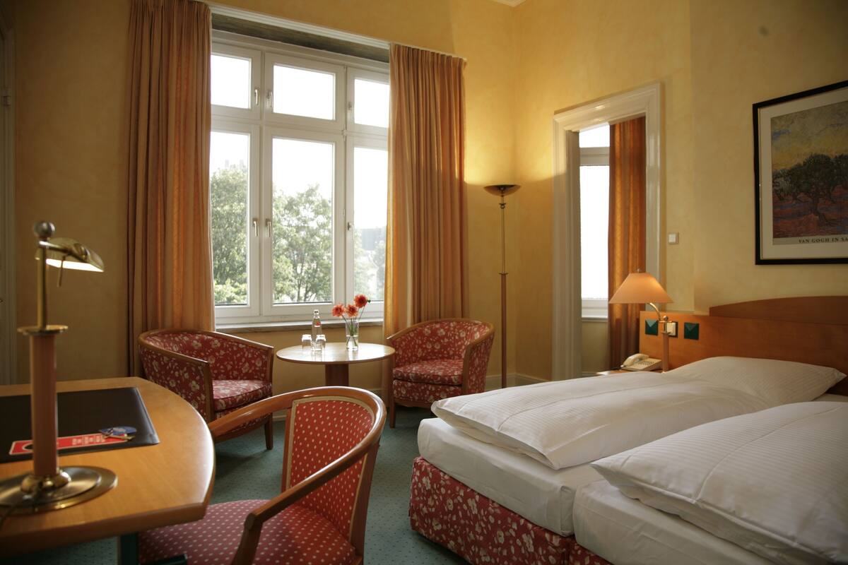 Habitación de hotel con dos camas, un escritorio con lámpara, una silla, una mesita de noche con un jarrón de flores y una ventana con cortinas.