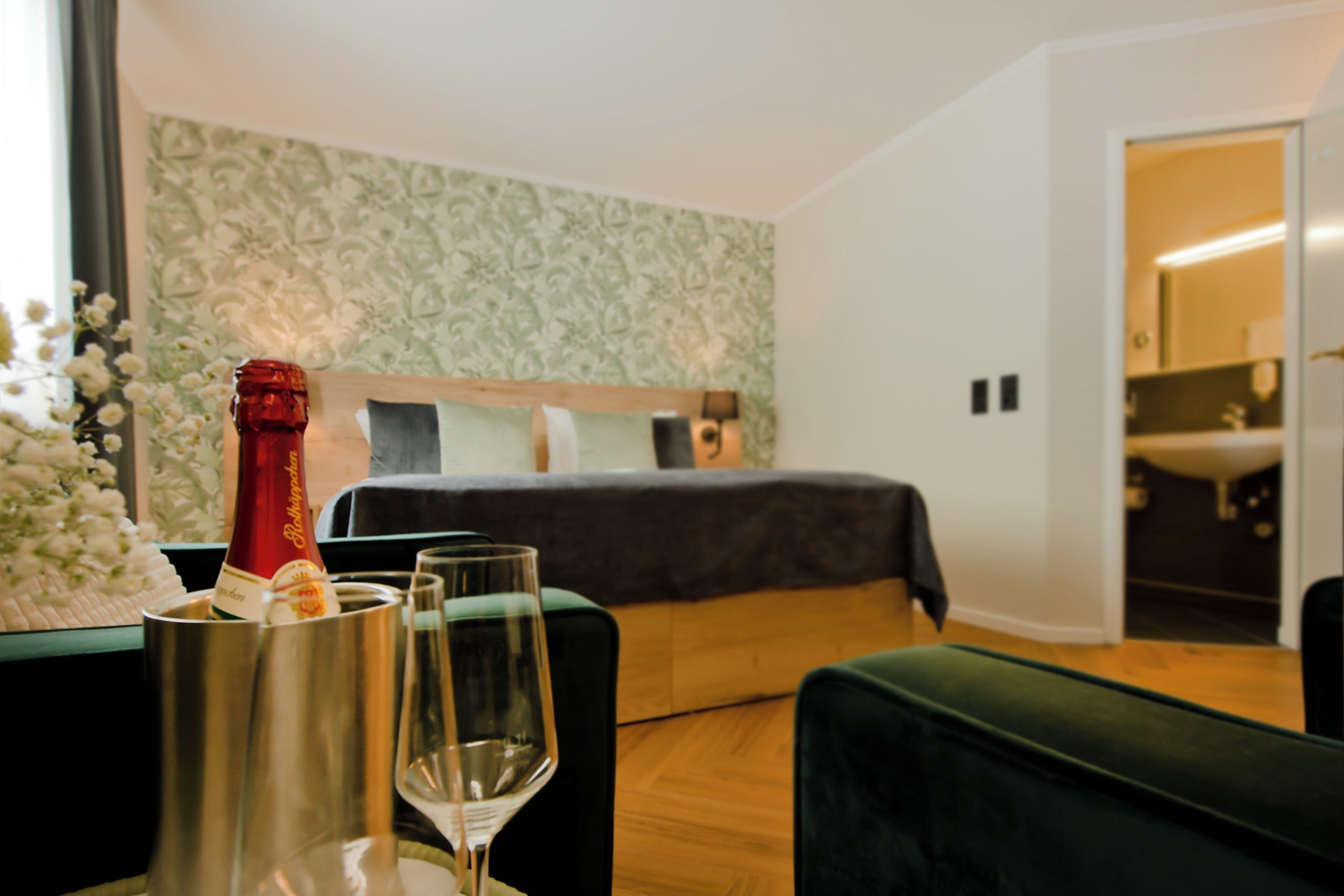 Habitación de hotel con cama, un sillón verde, una mesita con una botella y vasos, y un papel tapiz estampado.