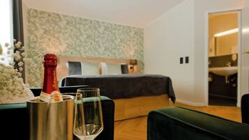 Habitación de hotel con cama, un sillón verde, una mesita con una botella y vasos, y un papel tapiz estampado.